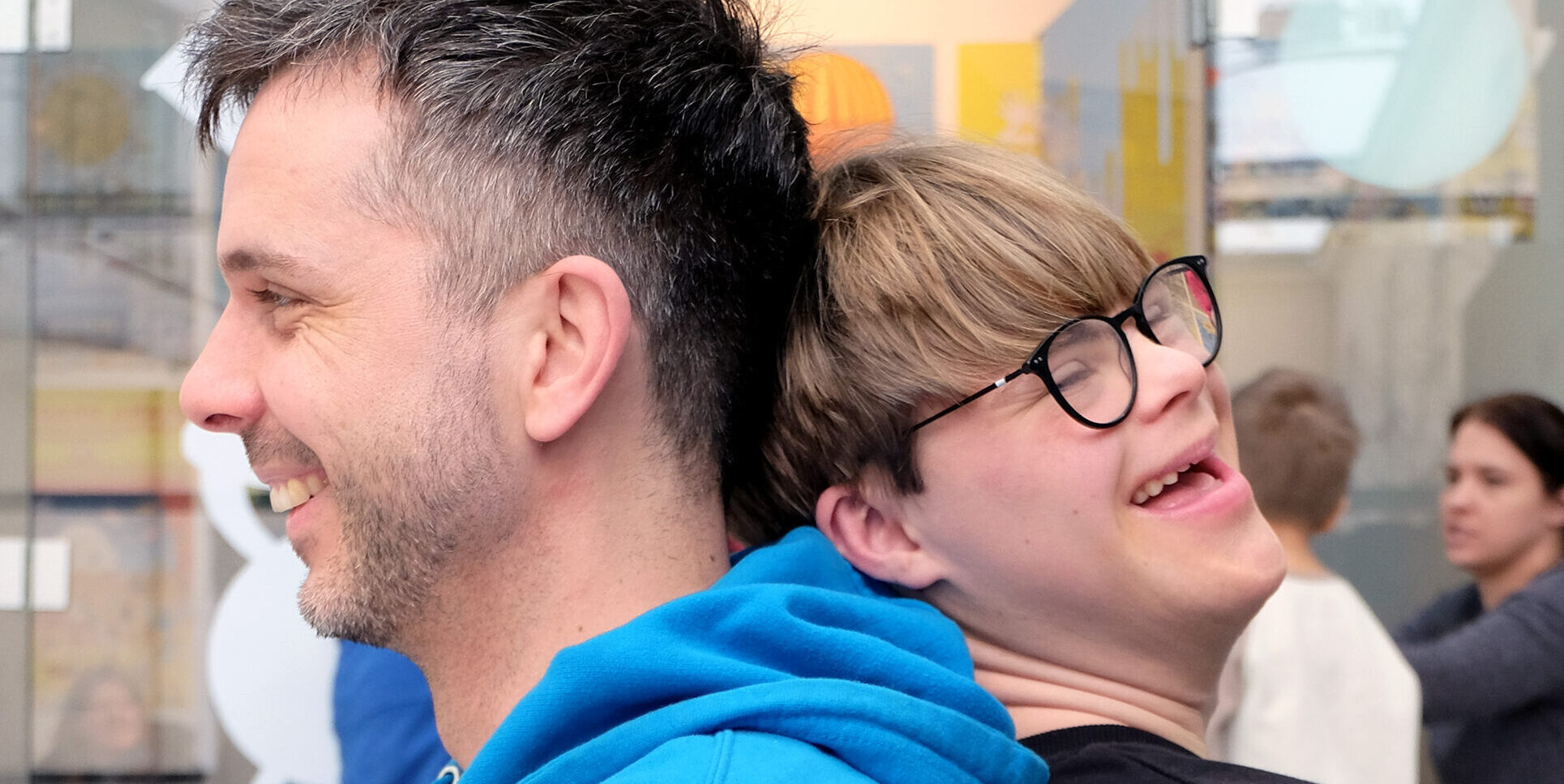 Ein erwachsener Mann mit dunklem Haar und Dreitagebart trägt einen blauen Hoodie. Er steht Rücken an Rücken mit einem Jugendlichen mit blonder Brille und schwarzem Pullover. Beide lachen und wirken fröhlich. Im Hintergrund befindet sich ein moderner, lichtdurchfluteter Raum mit Glaswänden, in dem weitere Personen zu sehen sind.